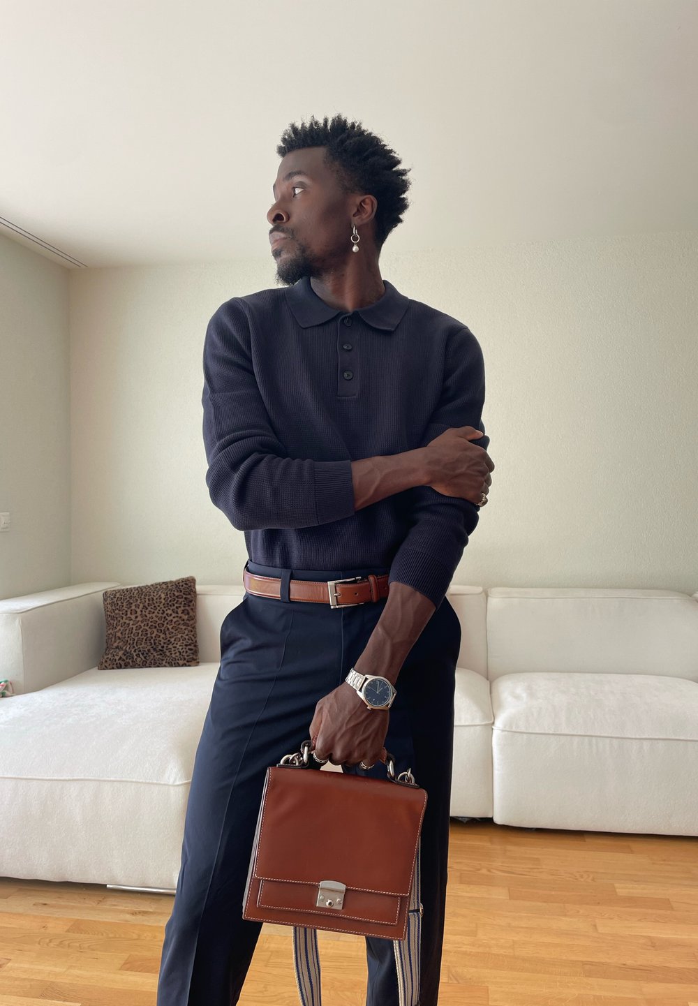 Mann in marineblauem Pullover und Hose, der eine braune Ledertasche hält und in einem Raum mit weißem Sofa und Holzboden steht.