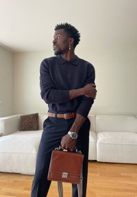 Mann in marineblauem Pullover und Hose, der eine braune Ledertasche hält und in einem Raum mit weißem Sofa und Holzboden steht.