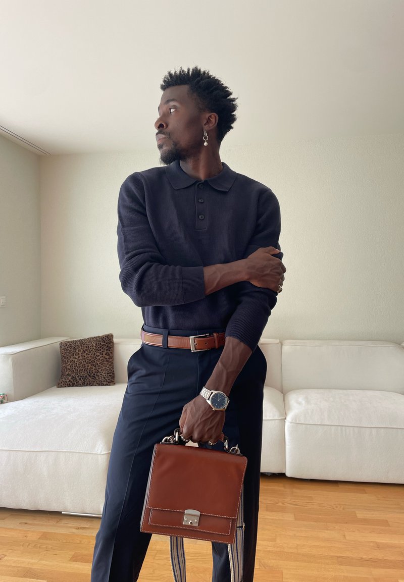 Bărbat îmbrăcat în pulover albastru închis și pantaloni, ținând o geantă maro de piele, stând într-o cameră cu canapea albă și podea de lemn.