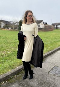 Beżowa prążkowana sukienka z czarną lamówką, zestawiona z czarną kurtką ze sztucznego futra. Strój uzupełniają czarne botki i rajstopy. Tło z zieloną trawą.