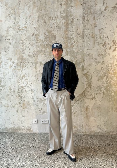 Chaqueta de cuero negra, camisa a rayas azules, corbata con estampado, pantalones anchos beige y zapatos negros con acentos blancos. Fondo neutral.