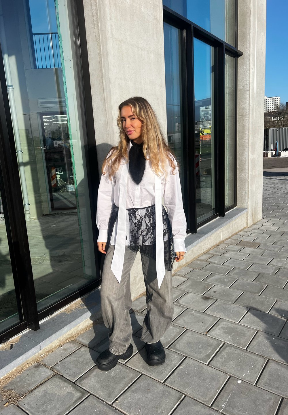 Une femme aux longs cheveux blonds se tient sur un trottoir à côté d'un bâtiment en verre, portant un pantalon gris ample, une chemise blanche avec un détail en dentelle noire et des chaussures noires.