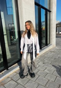 Une femme aux longs cheveux blonds se tient sur un trottoir à côté d'un bâtiment en verre, portant un pantalon gris ample, une chemise blanche avec un détail en dentelle noire et des chaussures noires.