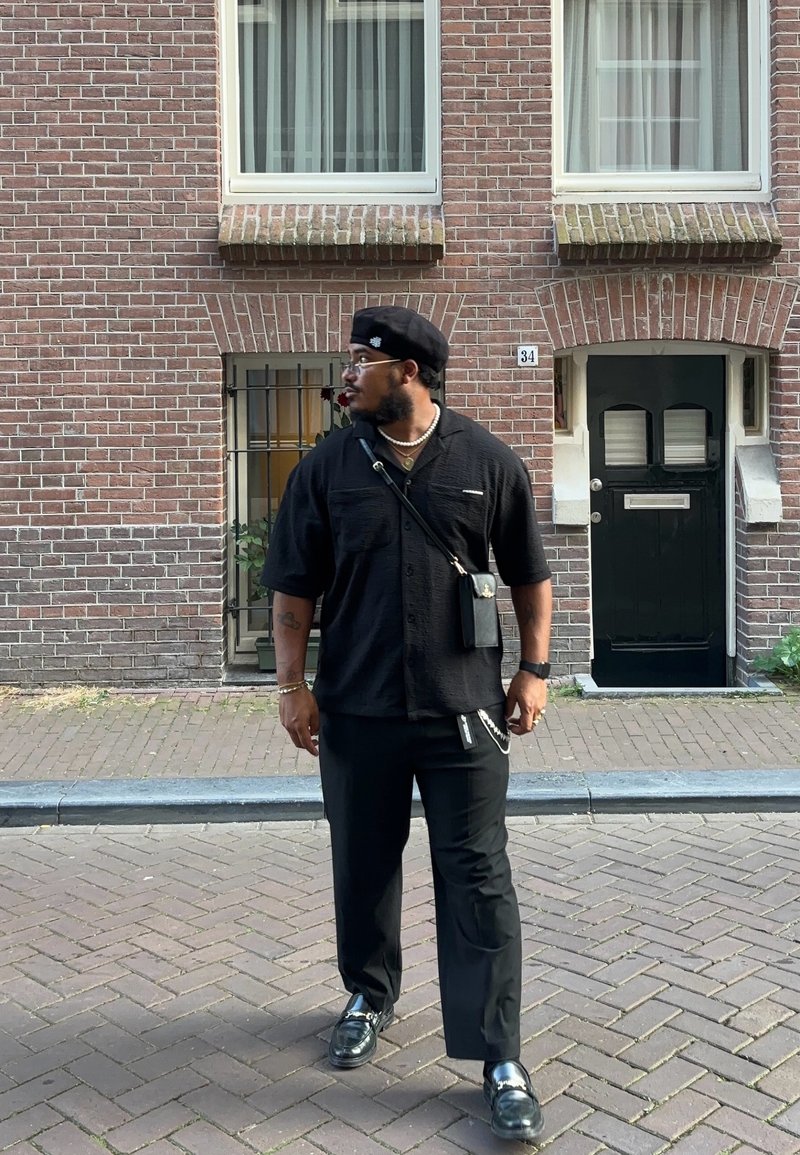 Μαύρο πουκαμισάκι με κοντά μανίκια, μαύρο παντελόνι, γυαλιστερά μαύρα παπούτσια και μπερέ; κρατώντας μια μικρή τσάντα; αστικός τοίχος από τούβλα.