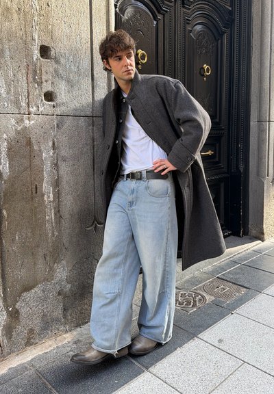 Joven con abrigo gris, camiseta blanca, vaqueros claros holgados y botas apoyado en una pared de piedra junto a una puerta negra ornamentada.