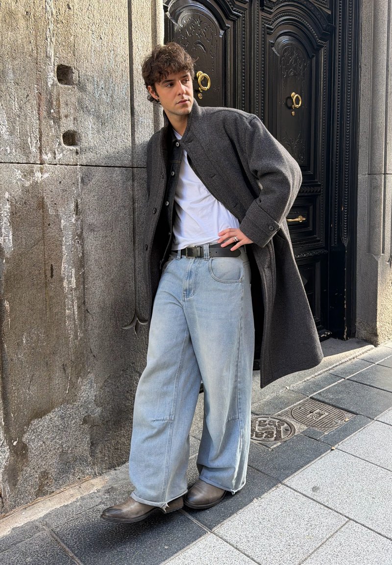 Ung mann i grå kåpe, hvit T-skjorte, vide lyse jeans og støvler som lener seg mot en steinvegg ved siden av en svart, dekorativ dør.