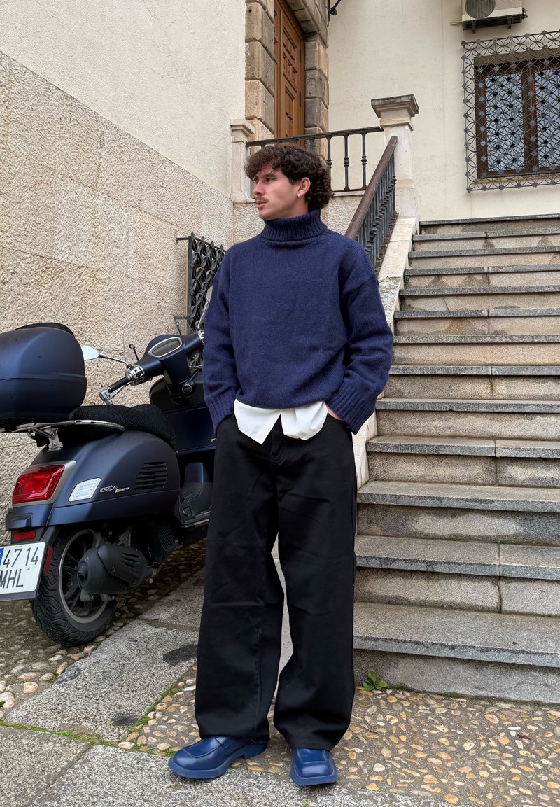 Pull à col roulé en maille bleu marine, superposé sur une chemise blanche, associé à un pantalon large noir et des chaussures bleu foncé à enfiler, debout à côté d'un scooter.