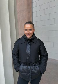 Czarna kurtka puchowa o błyszczącym wykończeniu, z paskiem w talii i pikowanym wzorem. Posiada szeroki kołnierz i zamek błyskawiczny.