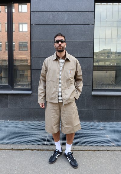 Hombre con chaqueta beige y pantalones cortos, camisa de rayas, gafas de sol negras, calcetines blancos y zapatillas negras Adidas, de pie junto a una pared oscura con azulejos.