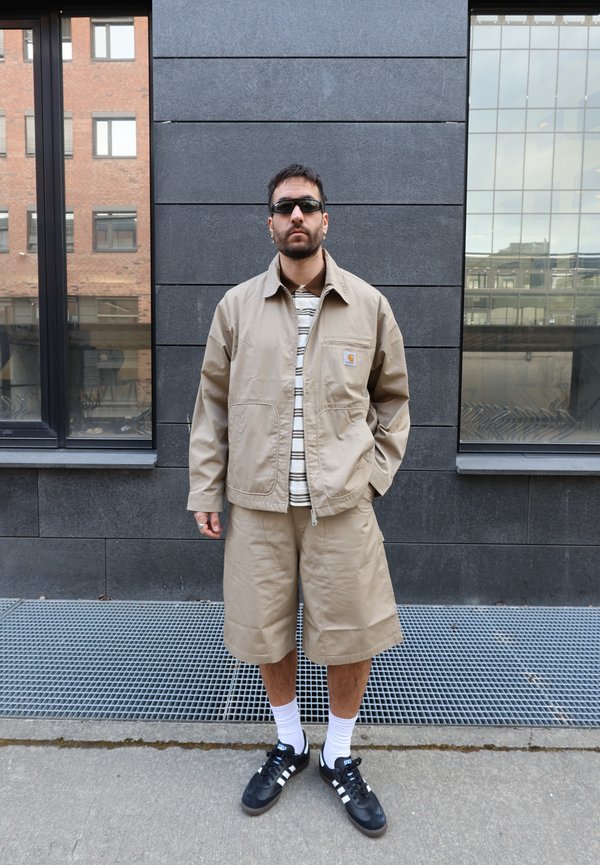 Homme portant une veste beige et un short, une chemise à rayures, des lunettes de soleil noires, des chaussettes blanches et des baskets Adidas noires, debout contre un mur carrelé sombre.