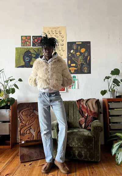 Una persona con gafas y una chaqueta de pelaje está de pie en una habitación con suelo de madera, un sillón verde, plantas y varios carteles en la pared.