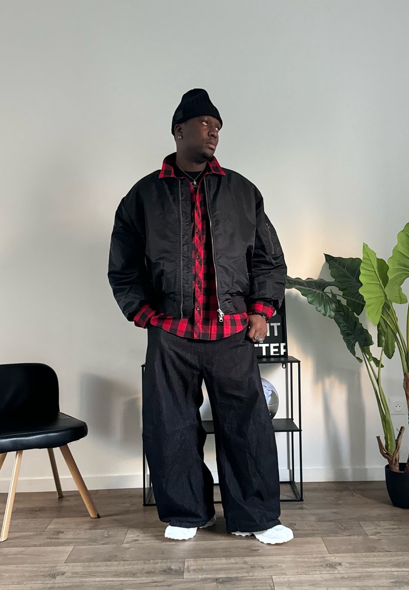 Veste bomber noire sur une chemise à carreaux rouge et noir, associée à un pantalon large noir et des chaussures blanches. Cadre intérieur avec des plantes.