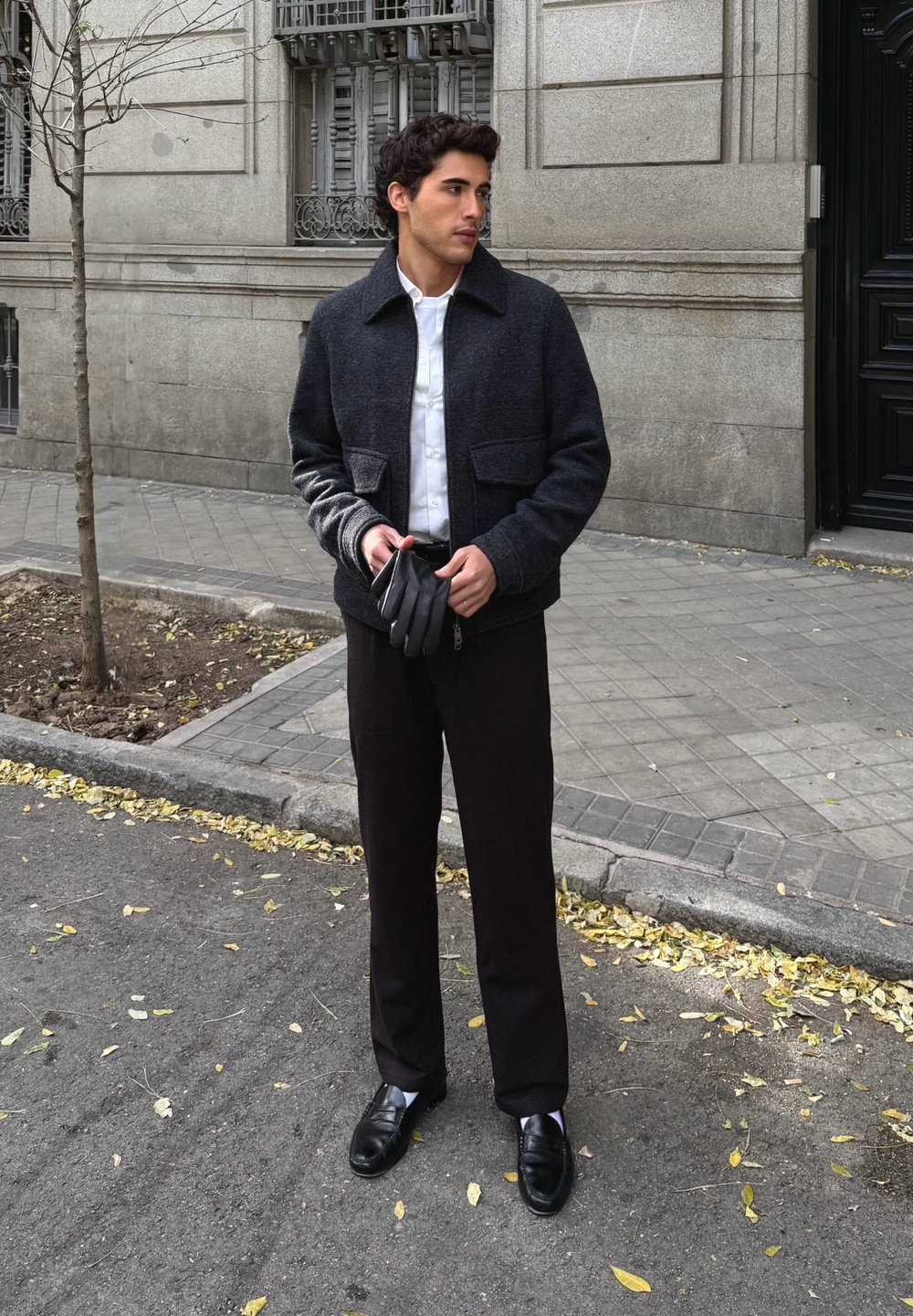 Giacca scura in misto lana con collo a punta, camicia bianca abbottonata, pantaloni neri e scarpe nere strette. Tiene in mano guanti in pelle nera.