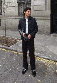 Giacca scura in misto lana con collo a punta, camicia bianca abbottonata, pantaloni neri e scarpe nere strette. Tiene in mano guanti in pelle nera.