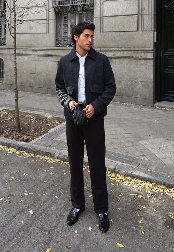 Tume villa segust jope laia kraega, valge nööpidega särgiga, mustade pükste ja kitsaste mustade kingadega. Hoides musti nahkkindaid.