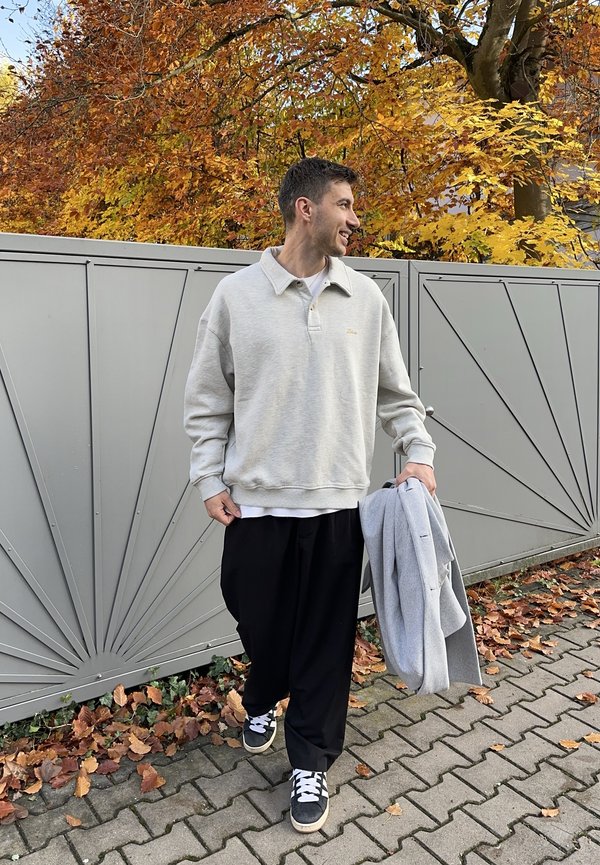 Light gray fleece pullover with a collar, gold accent, black loose-fitting trousers, and black sneakers with white laces, held gray jacket.