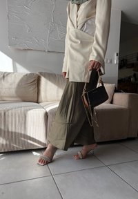 Άτομο που φοράει ελαιόχρωμα παντελόνια φαρδιάς γραμμής, κρεμ σακάκι και ψηλοτάκουνα με διαφανείς ιμάντες, κρατώντας μαύρη τσάντα χειρός με χρυσή αλυσίδα σε ένα φωτε ινό σαλόνι.