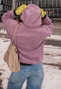 Personne portant une polaire à capuche violette avec un motif tourbillonnant et "WIP" au dos, des gants jaunes, un jean bleu, et portant un sac à bandoulière beige dans une zone urbaine enneigée.