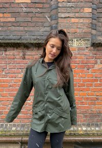 Giacca verde impermeabile con cappuccio, bottoni a pressione e due tasche anteriori. Abbinata a leggings neri di fronte a una parete di mattoni.