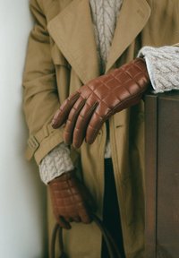 Bruna kviltade läderhandskar med en slät yta och anpassad design. Bärs av en person i en beige trenchcoat, som visar upp eleganta detaljer.