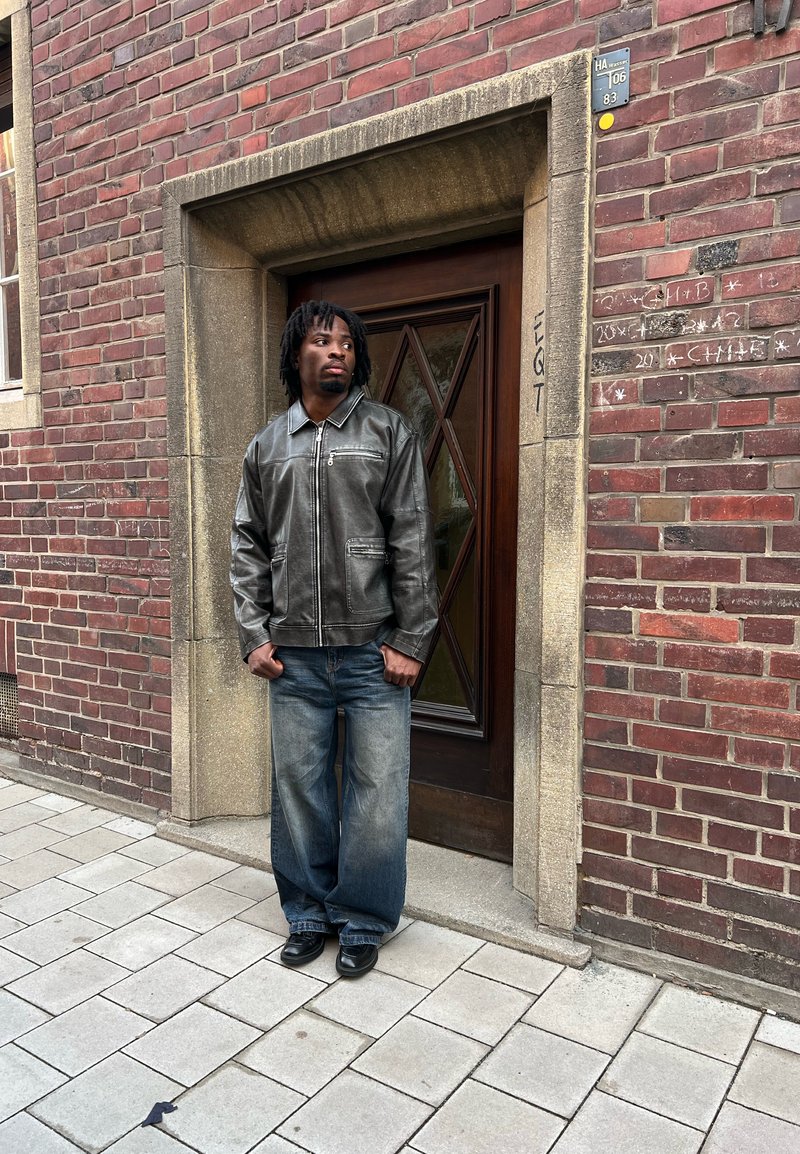 Veste en cuir noir avec poches zippées, associée à un jean large en denim bleu et des chaussures noires, sur fond de mur en briques et porte en bois.