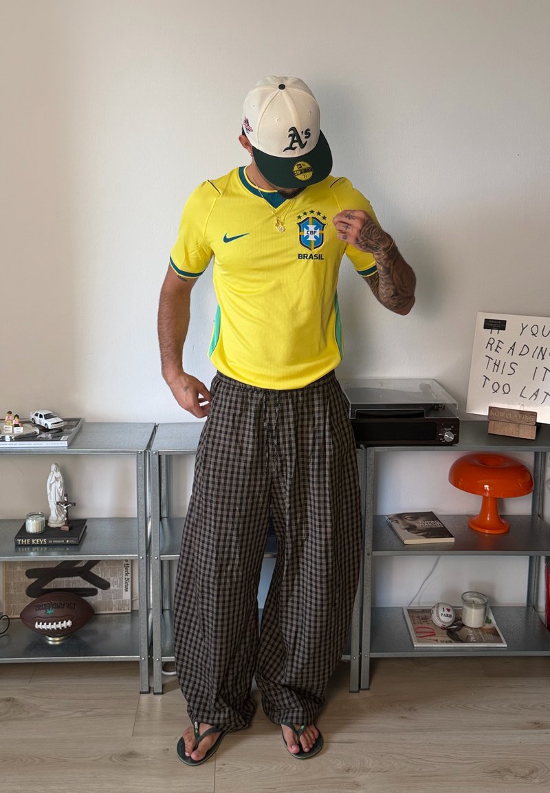 Homme portant un maillot de football jaune du Brésil, un pantalon ample à carreaux, une casquette de baseball blanche et verte, et des tongs, debout dans une pièce avec des étagères à proximité.