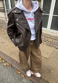 Geacă maro din piele de motociclist cu buzunare cu fermoar, hanorac gri și pantaloni kaki largi. Pantofi sport albi cu accente roșii completează ținuta.