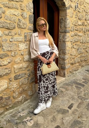 Camiseta sin mangas blanca, camisa beige de corte oversized, falda larga estampada en marrón y blanco, y zapatillas blancas. Sosteniendo un bolso tejido en crema con un acento marrón.