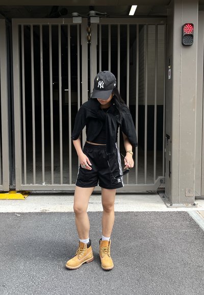 Top corto negro y pantalones cortos, botas de trabajo color tostado con acentos negros, sosteniendo gafas de sol. De pie frente a una entrada con reja.