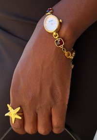 Orologio da polso dorato con quadrante bianco e gemme decorative rosse e ambra. Un anello a forma di stella marina con texture abbellisce la mano, arricchendo l'insieme.