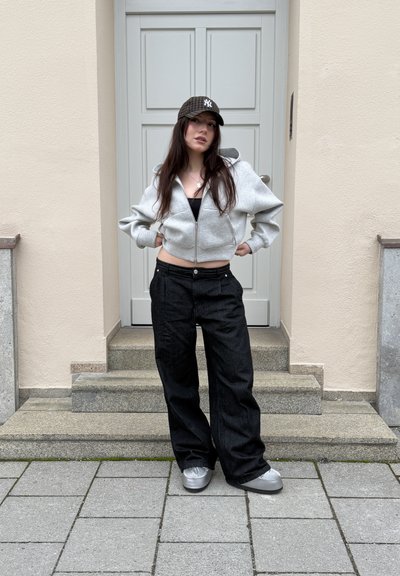 Joven con cabello largo que lleva una sudadera gris con cremallera, pantalones negros de pierna ancha, zapatos plateados y una gorra de béisbol a cuadros oscuros, de pie sobre unas escaleras.