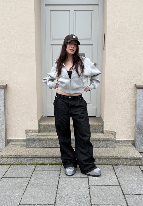 Jonge vrouw met lang haar, gekleed in een grijze hoodie met rits, zwarte wijde broek, zilveren schoenen en een donker geruit baseballpet, staande op trappen.