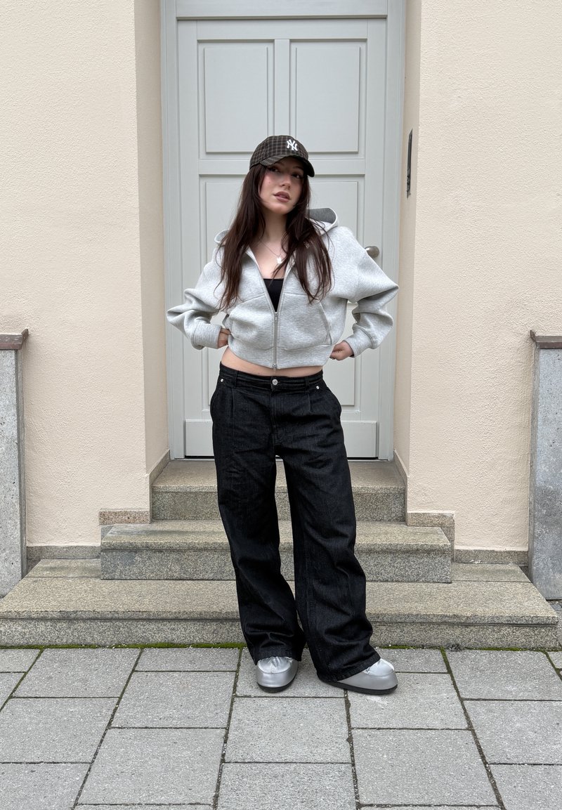 Tânără cu păr lung, purtând o hanorac gri cu fermoar, pantaloni negri largi, pantofi argintii și o șapcă de baseball cu model în carouri închise, stând pe trepte.