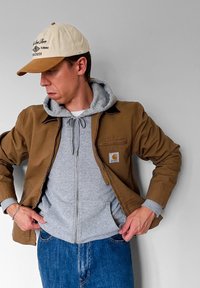 Hnědá pracovní bunda přes šedou mikinu s kapsou na zip a klokankou, doplněná modrými džíny a béžovou čepicí.