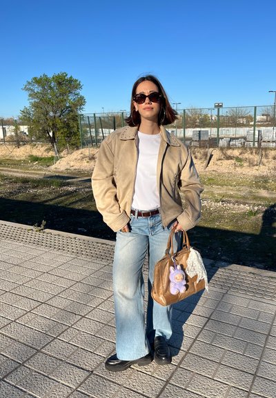 Chaqueta de ante beige con acentos de tachuelas, camiseta blanca, jeans acampanados de color azul claro, bolso marrón con un charm de peluche púrpura, zapatos negros.