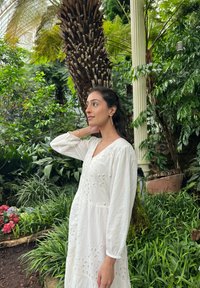 Robe blanche à manches longues avec broderie en œillets et décolleté en V. Portée dans un environnement luxuriant rempli de verdure et de plantes tropicales.