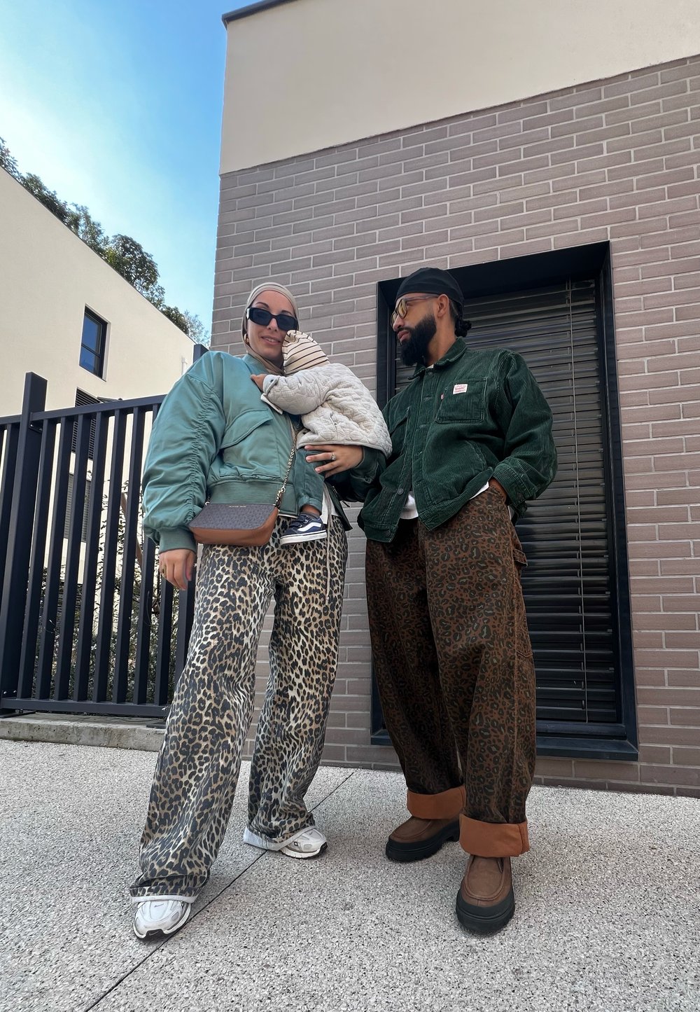 Δύο άτομα στέκονται έξω, το ένα φορούσε ένα τιρκουάζ μπόμπερ και παντελόνι με λεοπάρ σχέδιο, και το άλλο ένα πράσινο τζάκετ από καμπαρντίνα και καφέ παντελόνι με σχέδιο.