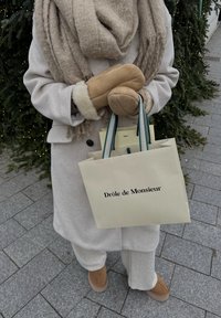 Personne en manteau beige, écharpe et moufles tenant des sacs de shopping portant l'inscription « Drôle de Monsieur » près d'arbres à feuilles persistantes décorés sur un trottoir pavé.