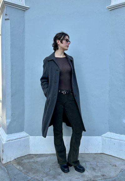 Abrigo largo gris de tela texturada, usado sobre una blusa marrón y pantalones de pana verde oscuro. Botines negros completan el conjunto.