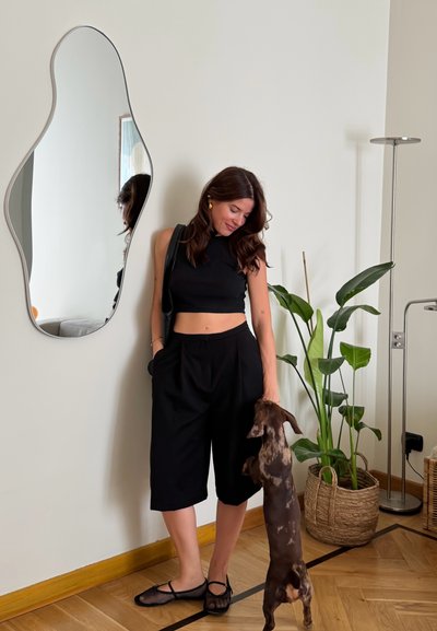 Top corto negro y culottes negros de corte holgado. Zapatos planos de malla negros. Cabello ondulado. Reflejada en un espejo ondulado. Plantas verdes y una cesta cerca.