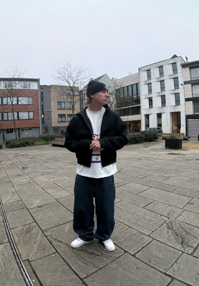 Sudadera negra con cremallera sobre una camisa blanca, combinada con jeans azules holgados y zapatillas blancas. El entorno presenta arquitectura moderna.