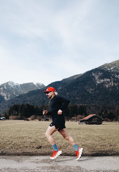 Corredor con una camiseta de manga larga negra y pantalones cortos negros, gorra roja, calcetines azules y zapatos blanco-rojos, en un camino de grava con montañas al fondo.