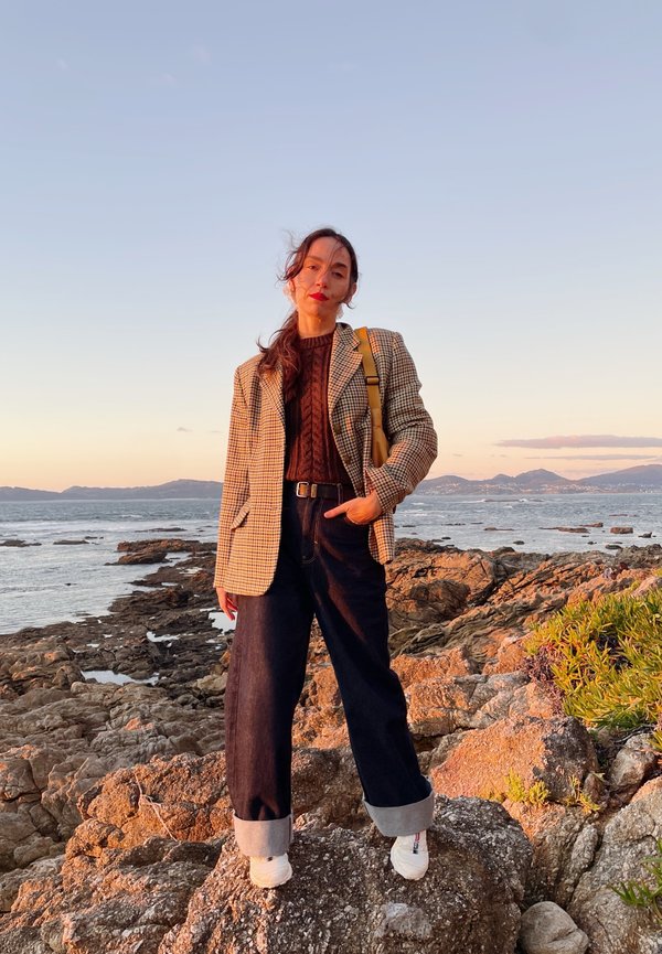 Karierte Blazer über einem braunen Pullover, hochgeschnittene dunkle Jeans mit umgeschlagenen Säumen und weiße Sneakers, vor einer felsigen Küstenlandschaft.