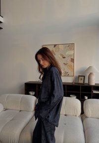 Temno modra vrhnja tkanina z teksturo in hlače z ohlapnimi rokavi, ki jih nosi oseba ob svetloživi, šivani zofe in abstraktni stenski umetnini.