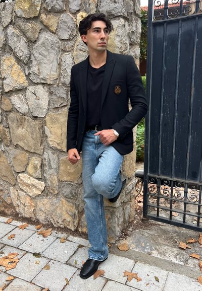 Blazer negro con emblema de escudo, camiseta negra, jeans azul claro y zapatos negros. Está de pie contra una pared de piedra con hojas en el suelo.