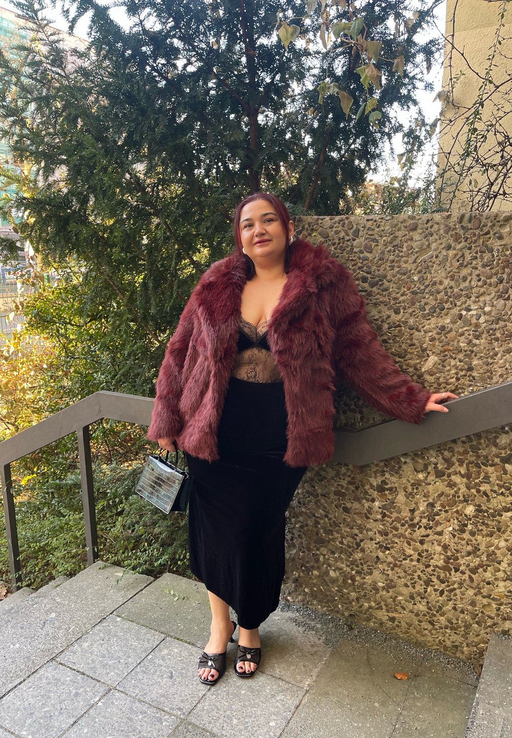 Manteau en fausse fourrure bordeaux, body en dentelle noire, jupe en velours noire et mules noires. Petit sac noir à texture crocodile. Cadre extérieur.