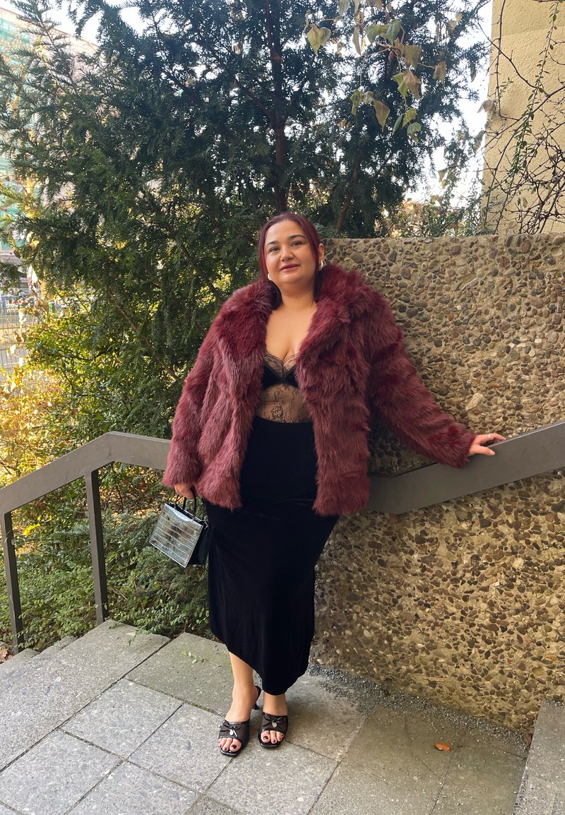 Manteau en fausse fourrure bordeaux, body en dentelle noire, jupe en velours noire et mules noires. Petit sac noir à texture crocodile. Cadre extérieur.