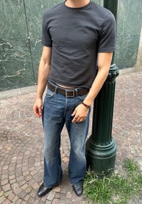 Person iført en mørkegrå t-shirt, løse blå jeans og sorte sko, stående ved siden af en grøn gadelampe på en brostensbelagt fortov.