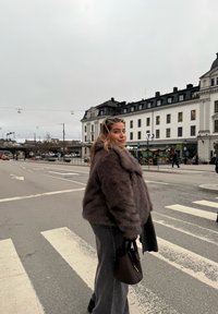 Ung kvinna i en brun pälsjacka som bär en mörk handväska, står på ett övergångsställe i en europeisk stad med historiska byggnader och molnig himmel.