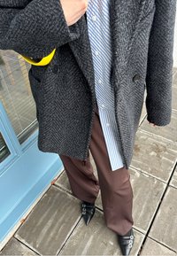 Γκρι παλτό σε σχέδιο ρόμβου πάνω από μπλε και λευκό ριγέ πουκάμισο, συνδυασμένο με καφέ παντελόνι και μυτερά μαύρα παπούτσια με διπλά μπουτόν.